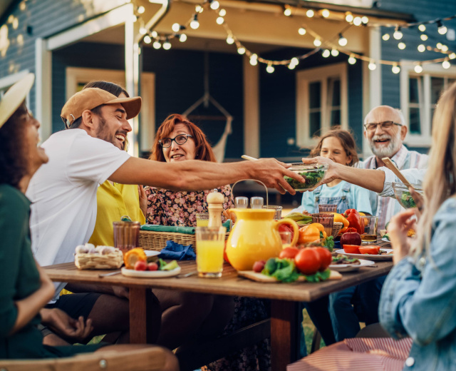 Foto einer mehrgenerationalen Familie, die draußen um einen Esstisch sitzt. Die Menschen lachen. Auf dem Tisch stehen Schalen mit Tomaten und Paprika. Ein Mann in der Mitte beugt sich über den Tisch und nimmt eine Salatschale von einer anderen Person entgegen. Im Hintergrund ist eine leuchtende Lichterkette zu sehen.
